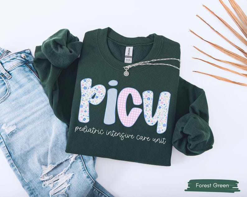 May include: Forest green sweatshirt with the letters "PICU" in a floral and gingham pattern. The words "pediatric intensive care unit" are printed below the letters. A silver necklace is draped across the sweatshirt, which is laid on top of a pair of blue jeans.