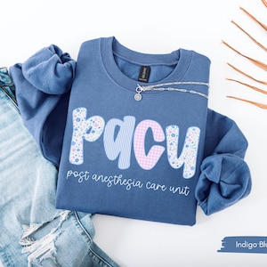 May include: Indigo blue sweatshirt with the letters "PACU" in a floral and gingham pattern. The text "post anesthesia care unit" is below the letters. A silver necklace is on the sweatshirt, and a pair of blue jeans are next to it.