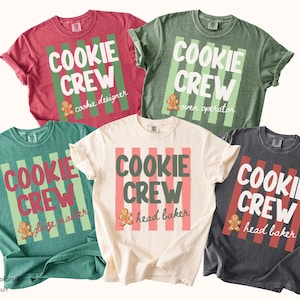 May include: Five short-sleeve t-shirts in red, olive green, teal, cream, and charcoal gray. Each shirt features a vertical striped background, the text COOKIE CREW, and a small gingerbread person graphic with specific titles like cookie designer, oven operator, glaze master, and head baker.