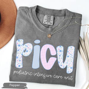 Puede incluir: Una camiseta gris con la palabra "PICU" en un dise&ntilde;o floral y de cuadros. El texto "pediatric intensive care unit" est&aacute; debajo de la palabra PICU. Un collar dorado est&aacute; drapeado sobre la camiseta.