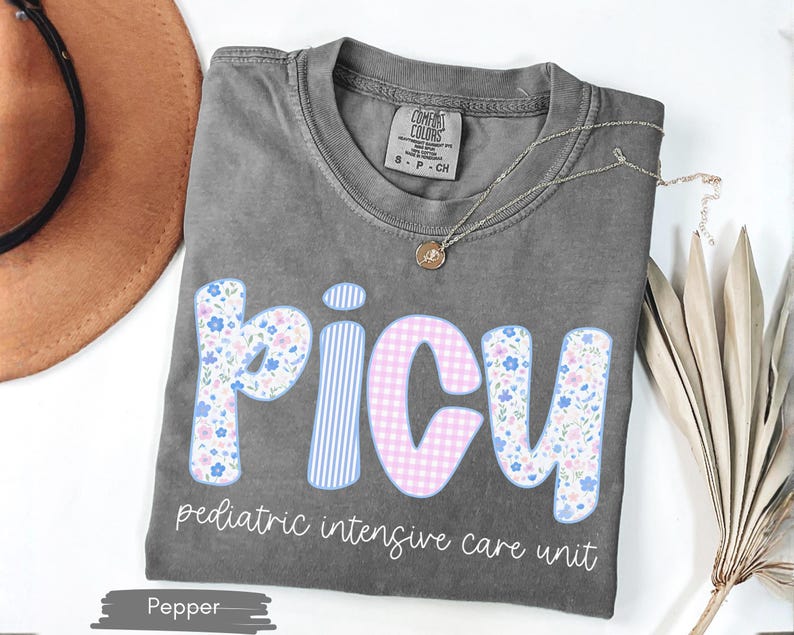 Puede incluir: Una camiseta gris con la palabra "PICU" en un dise&ntilde;o floral y de cuadros. El texto "pediatric intensive care unit" est&aacute; debajo de la palabra PICU. Un collar dorado est&aacute; drapeado sobre la camiseta.