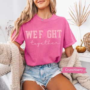 K&ouml;nnte beinhalten: Ein leuchtend pinkfarbenes T-Shirt mit dem Aufdruck "WEF GHT together" in Hellrosa. Das Shirt wird mit Jeansshorts kombiniert. Ein beigefarbener Cardigan ist &uuml;ber die Schultern gelegt.