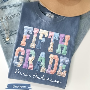 May include: A blue jean-colored t-shirt with the words "FIFTH GRADE" in colorful patchwork letters. The name "Mrs. Anderson" is written below the text. A tan hat and blue jeans are also in the image.