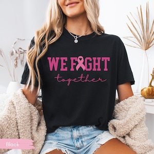 K&ouml;nnte beinhalten: Schwarzes T-Shirt mit dem Text "WE FIGHT together" in Pink, mit einer rosa Schleifengrafik. Das Shirt ist ein klassischer Rundhalsausschnitt.