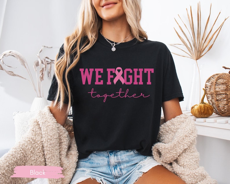 K&ouml;nnte beinhalten: Schwarzes T-Shirt mit dem Text "WE FIGHT together" in Pink, mit einer rosa Schleifengrafik. Das Shirt ist ein klassischer Rundhalsausschnitt.