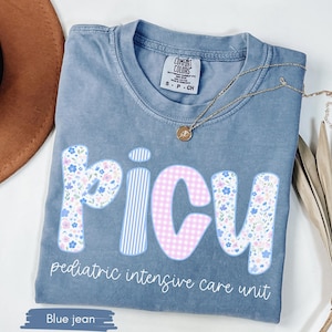 May include: Blue jean Comfort Colors t-shirt with "PICU" in floral and gingham letters, and "pediatric intensive care unit" printed below. A gold necklace with a pendant rests on the shirt.