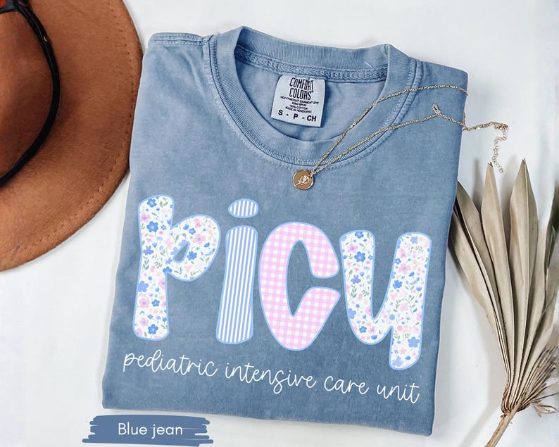 Puede incluir: Camiseta azul vaquero Comfort Colors con "PICU" en letras florales y de cuadros, y "pediatric intensive care unit" impreso debajo. Un collar de oro con un colgante descansa sobre la camisa.