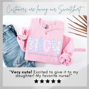 Op de afbeelding: Lichtroze sweatshirt met de letters "NICU" in bloemen- en ruitpatroon. De tekst "NICU neonatal intensive care unit" staat op de voorkant. De afbeelding bevat ook een spijkerbroek en een gouden ketting.
