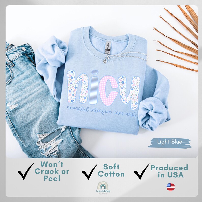 Op de afbeelding: Lichtblauw NICU-sweatshirt met bloemen- en ruitletters. De tekst "neonatal intensive care unit" staat eronder. Het sweatshirt wordt getoond met een blauwe jeans. Gemaakt van zacht katoen en geproduceerd in de VS.