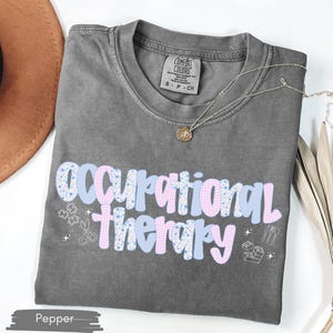 Op de afbeelding: Een grijze t-shirt met de tekst "Occupational Therapy" in een speels lettertype, versierd met lichtblauwe en roze patronen. Het shirt bevat kleine afbeeldingen van scharen, puzzelstukjes en andere therapie-gerelateerde symbolen. Een gouden ketting ligt op het shirt.