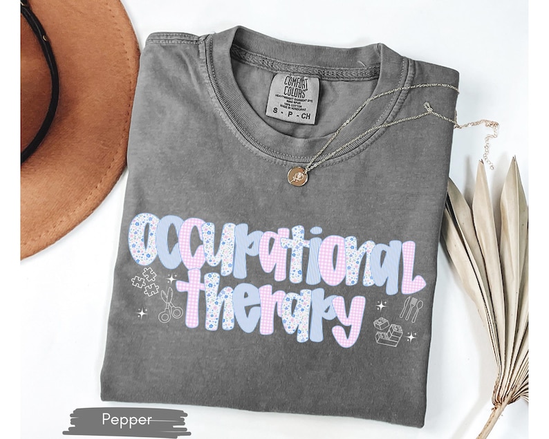 Op de afbeelding: Een grijze t-shirt met de tekst "Occupational Therapy" in een speels lettertype, versierd met lichtblauwe en roze patronen. Het shirt bevat kleine afbeeldingen van scharen, puzzelstukjes en andere therapie-gerelateerde symbolen. Een gouden ketting ligt op het shirt.