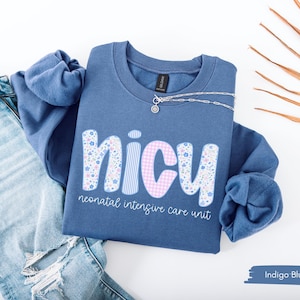 May include: Indigo blue sweatshirt with the word "NICU" in a floral and gingham pattern. The text "neonatal intensive care unit" is below the word "NICU". A silver necklace is on the sweatshirt, and a pair of ripped blue jeans are next to it.