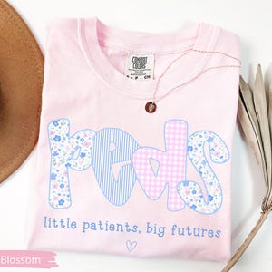 May include: A light pink t-shirt with the word "PEDS" in a playful font. The letters are decorated with blue stripes, floral patterns, and pink gingham. The text "little patients, big futures" is below the word "PEDS". A gold necklace is on the shirt.