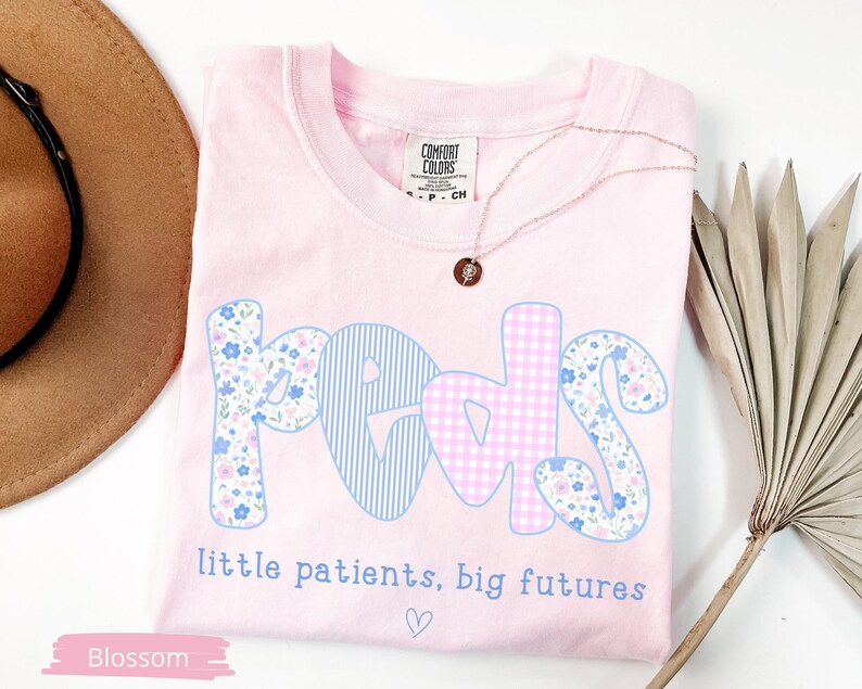 May include: A light pink t-shirt with the word "PEDS" in a playful font. The letters are decorated with blue stripes, floral patterns, and pink gingham. The text "little patients, big futures" is below the word "PEDS". A gold necklace is on the shirt.