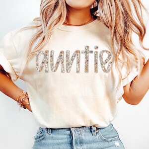May include: A cream-coloured t-shirt featuring the word "auntie" in a floral design. The letters are filled with small flowers in pink, purple, and green. The shirt has rolled-up sleeves and is worn with blue jeans.