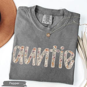May include: A grey Comfort Colors t-shirt featuring the word "AUNTIE" in a floral design. The shirt has a crew neck and short sleeves. A gold necklace with a pendant is on the shirt. A brown hat is in the background.