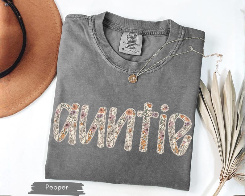 May include: A grey Comfort Colors t-shirt featuring the word "AUNTIE" in a floral design. The shirt has a crew neck and short sleeves. A gold necklace with a pendant is on the shirt. A brown hat is in the background.