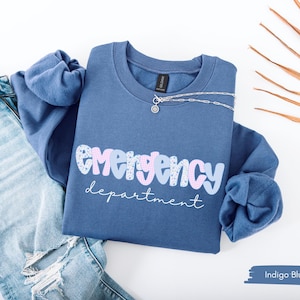 May include: Indigo blue crewneck sweatshirt with the words "emergency department" in a playful font. The word "emergency" is in a pastel color block design. A silver necklace rests on the sweatshirt, which is paired with light wash denim jeans.