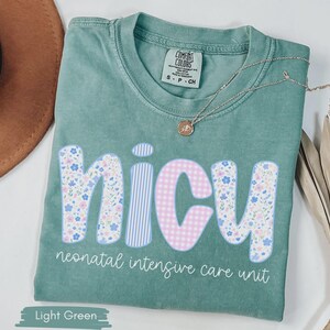Peut inclure: T-shirt vert clair avec le mot "NICU" dans un motif floral, ray&eacute; et vichy. Le texte "neonatal intensive care unit" est en dessous du mot. Un collier dor&eacute; avec un pendentif est sur le t-shirt.