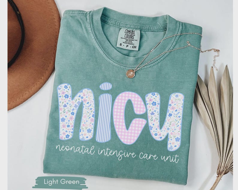 Peut inclure: T-shirt vert clair avec le mot "NICU" dans un motif floral, ray&eacute; et vichy. Le texte "neonatal intensive care unit" est en dessous du mot. Un collier dor&eacute; avec un pendentif est sur le t-shirt.