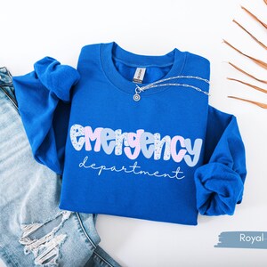 May include: Royal blue sweatshirt with the words "emergency department" in a playful font. The word "emergency" is in a bubble letter style with a mix of patterns and colors. A silver necklace is draped over the sweatshirt.