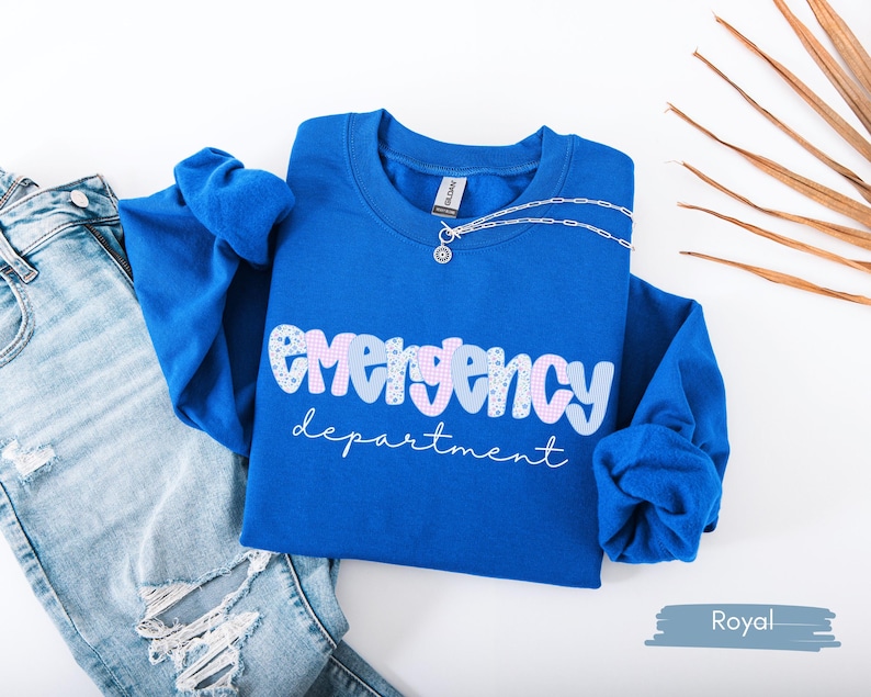 May include: Royal blue sweatshirt with the words "emergency department" in a playful font. The word "emergency" is in a bubble letter style with a mix of patterns and colors. A silver necklace is draped over the sweatshirt.