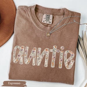 May include: Espresso-coloured t-shirt with the word "Auntie" embroidered in a floral design. The shirt has a crew neck and short sleeves. A silver necklace with a pendant is draped across the shirt. The shirt is made of a soft material.