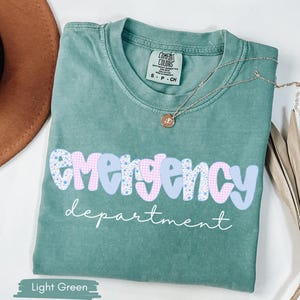 May include: Light green t-shirt with the words "emergency department" in a playful font. The word "emergency" is in a blue and pink gingham and floral pattern. A gold necklace with a pendant rests on the shirt.