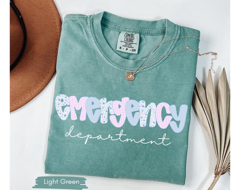 May include: Light green t-shirt with the words "emergency department" in a playful font. The word "emergency" is in a blue and pink gingham and floral pattern. A gold necklace with a pendant rests on the shirt.