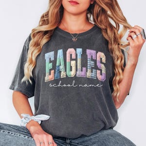 May include: Dark gray t-shirt with the word "EAGLES" in colorful patchwork letters. Below the word "EAGLES" is the phrase "school name" in a cursive font. The shirt is a casual style.