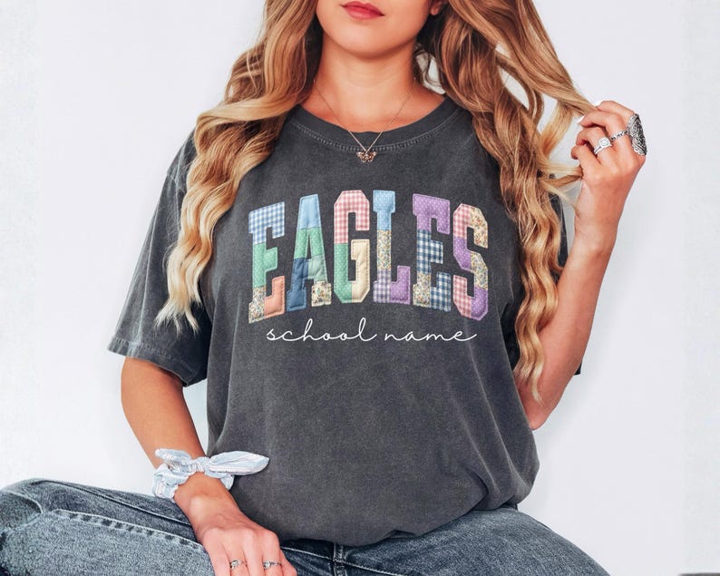 May include: Dark gray t-shirt with the word "EAGLES" in colorful patchwork letters. Below the word "EAGLES" is the phrase "school name" in a cursive font. The shirt is a casual style.