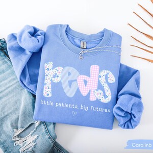 May include: Carolina blue sweatshirt with the word "peds" in a mix of floral, striped, and gingham patterns. The text "little patients, big futures" is below the word "peds". A silver necklace is draped on the sweatshirt. Light blue jeans are in the background.