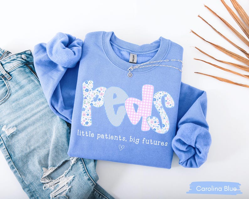 May include: Carolina blue sweatshirt with the word "peds" in a mix of floral, striped, and gingham patterns. The text "little patients, big futures" is below the word "peds". A silver necklace is draped on the sweatshirt. Light blue jeans are in the background.