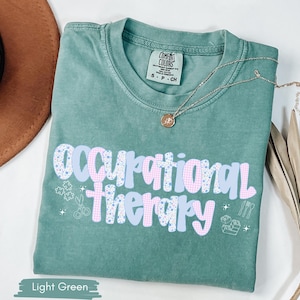 Op de afbeelding: Lichtgroen T-shirt met de woorden "Occupational Therapy" in een speels lettertype. De tekst is een mix van blauwe en roze patronen. Een gouden ketting met een hanger rust op het shirt. Het shirt heeft een Comfort Colors label.