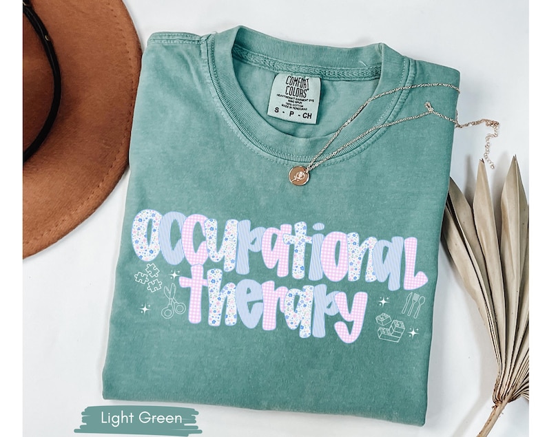 Op de afbeelding: Lichtgroen T-shirt met de woorden "Occupational Therapy" in een speels lettertype. De tekst is een mix van blauwe en roze patronen. Een gouden ketting met een hanger rust op het shirt. Het shirt heeft een Comfort Colors label.