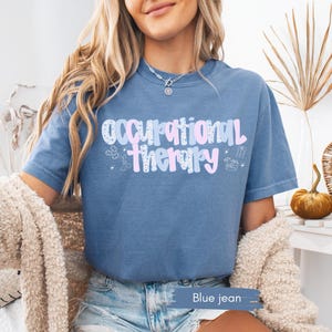 Op de afbeelding: Blauw spijkerstof T-shirt met korte mouwen met de tekst "Occupational Therapy" in een pastelkleurig ontwerp. Het shirt heeft een ronde hals en is gemaakt van een zacht materiaal. De tekst bevat kleine illustraties.