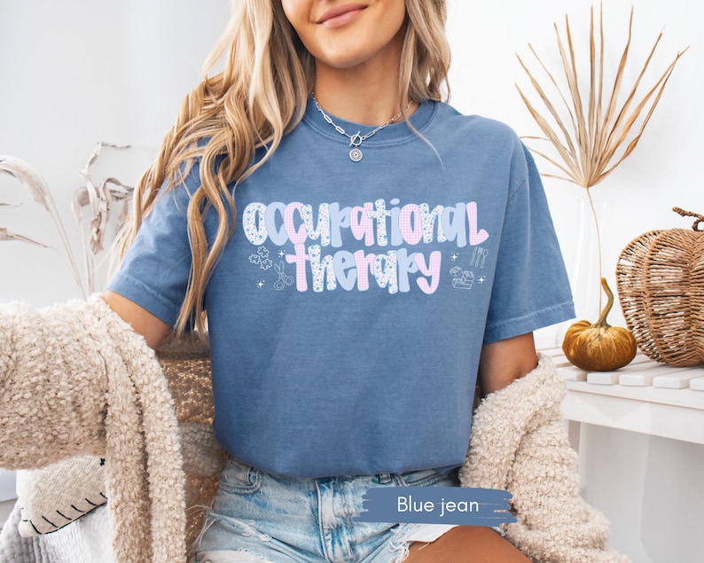 Op de afbeelding: Blauw spijkerstof T-shirt met korte mouwen met de tekst "Occupational Therapy" in een pastelkleurig ontwerp. Het shirt heeft een ronde hals en is gemaakt van een zacht materiaal. De tekst bevat kleine illustraties.