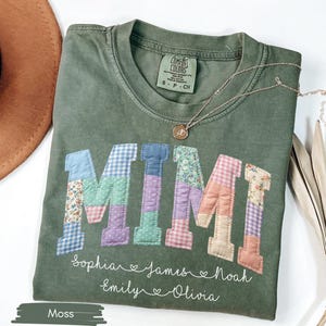 May include: Moss green t-shirt with the word "MIMI" in patchwork letters. Below the word are names: Sophia, James, Noah, Emily, and Olivia. A gold necklace rests on the shirt. The shirt is folded.