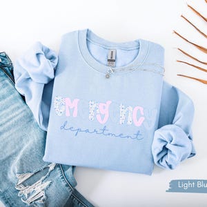 May include: Light blue sweatshirt with the text "EMERGENCY department" in a mix of pink and floral print. The sweatshirt is paired with light wash denim jeans. A silver necklace is draped on the sweatshirt.