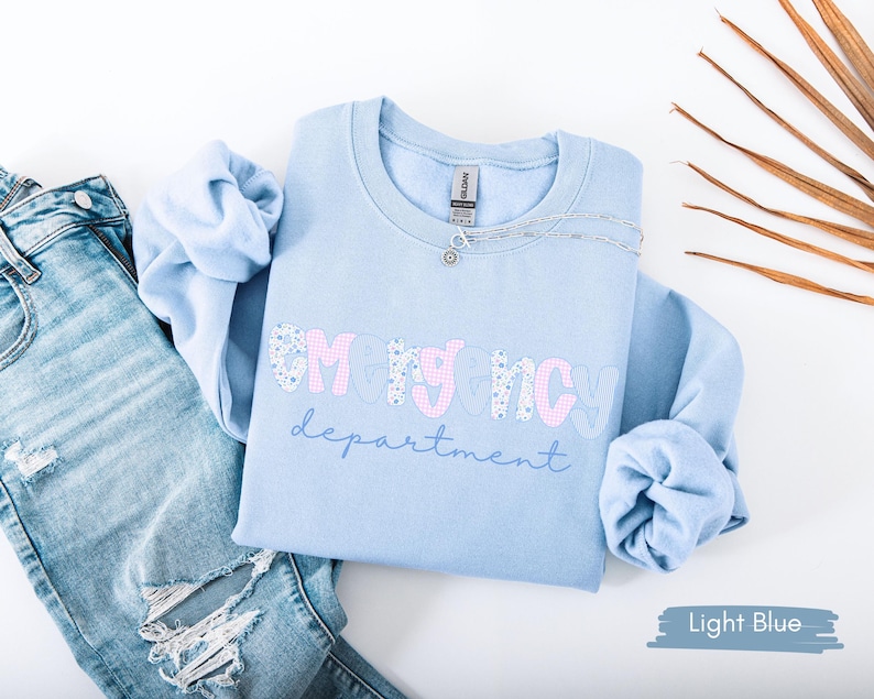 May include: Light blue sweatshirt with the text "EMERGENCY department" in a mix of pink and floral print. The sweatshirt is paired with light wash denim jeans. A silver necklace is draped on the sweatshirt.