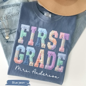 May include: A blue t-shirt with the words "FIRST GRADE" in colorful patchwork letters. The name "Mrs. Anderson" is written below the text. The shirt is lying on a pair of blue jeans, with a tan hat resting on top.