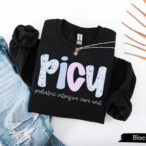 May include: Black PICU sweatshirt with floral and striped lettering, paired with light blue ripped jeans. The text "pediatric intensive care unit" is below the PICU letters. A silver necklace is draped across the sweatshirt.