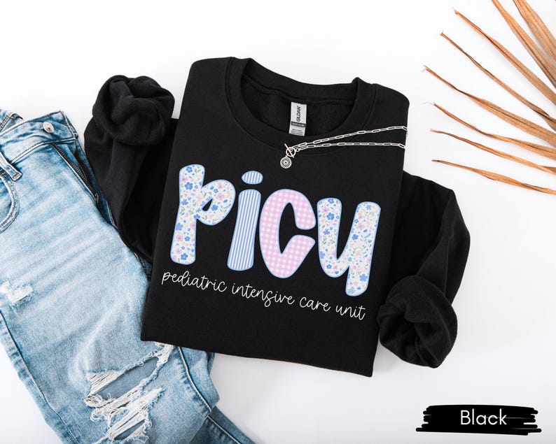 May include: Black PICU sweatshirt with floral and striped lettering, paired with light blue ripped jeans. The text "pediatric intensive care unit" is below the PICU letters. A silver necklace is draped across the sweatshirt.