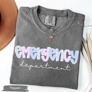 May include: A gray t-shirt with the words "emergency department" in a playful font. The word "emergency" is in a blue and pink patterned design. A gold necklace and a brown hat are also visible.