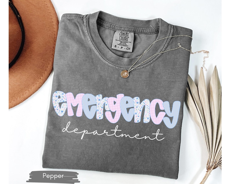 May include: A gray t-shirt with the words "emergency department" in a playful font. The word "emergency" is in a blue and pink patterned design. A gold necklace and a brown hat are also visible.
