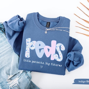 May include: Indigo blue sweatshirt with the word "peds" in a floral, striped, and gingham pattern. The text "little patients, big futures" is below the word "peds". A silver necklace and blue jeans are next to the sweatshirt.