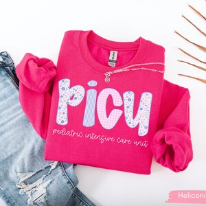 May include: A bright pink sweatshirt with the letters "PICU" in a floral and striped design. The text "pediatric intensive care unit" is below the letters. A silver necklace is draped across the shirt, which is next to a pair of blue jeans.