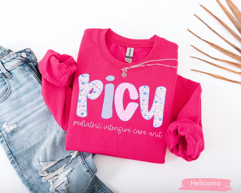 May include: A bright pink sweatshirt with the letters "PICU" in a floral and striped design. The text "pediatric intensive care unit" is below the letters. A silver necklace is draped across the shirt, which is next to a pair of blue jeans.