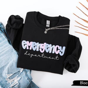 May include: Black crewneck sweatshirt with the words "emergency department" in a pastel design. A silver necklace rests on the sweatshirt, which is paired with light blue ripped jeans. The sweatshirt is a casual style.