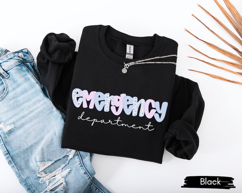 May include: Black crewneck sweatshirt with the words "emergency department" in a pastel design. A silver necklace rests on the sweatshirt, which is paired with light blue ripped jeans. The sweatshirt is a casual style.